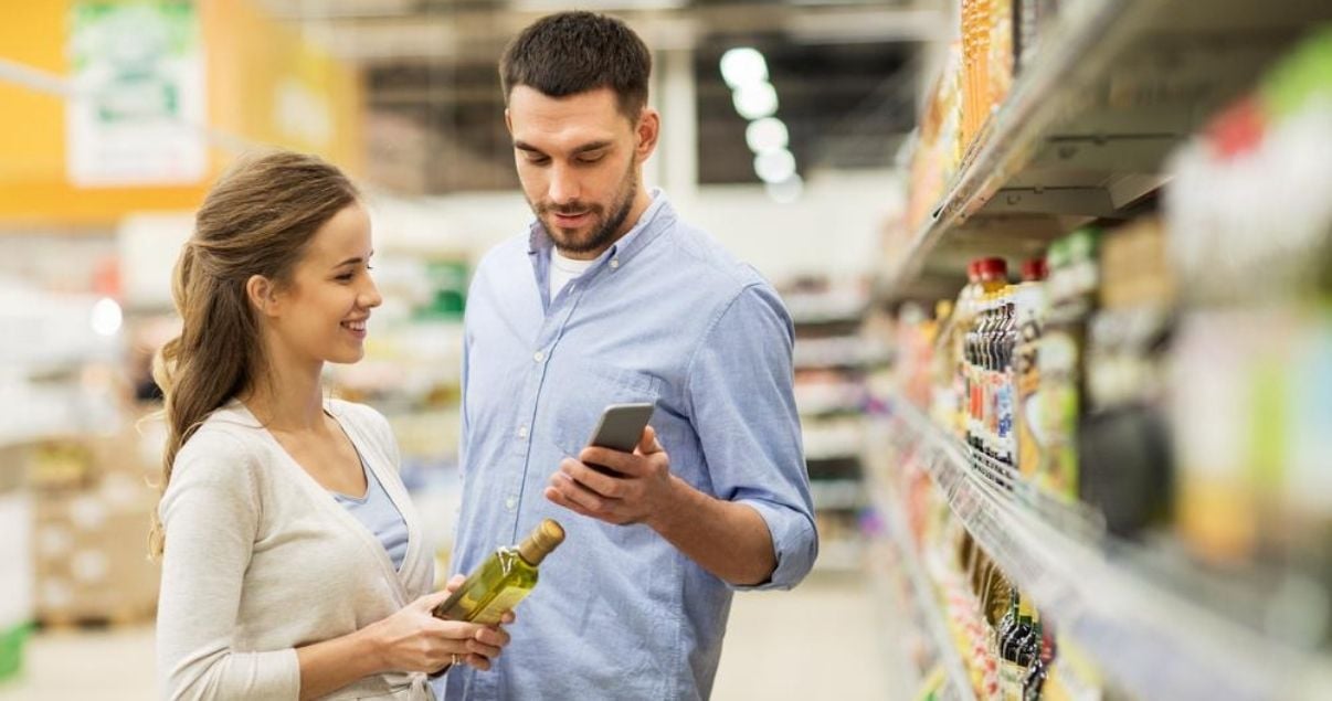 Pareja joven utilizando su celular en el supermercado con un producto que encontraron en la góndola gracias a la estrategia de facing